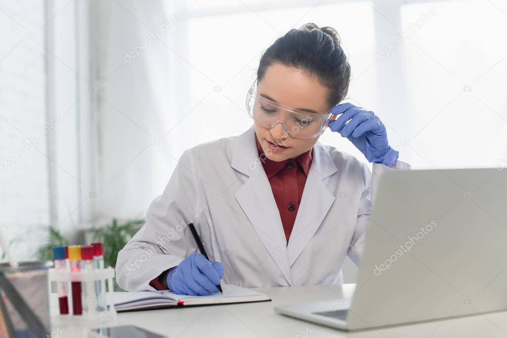 brunette scientist in latex gloves adjusting goggles while writing in notebook near devices and test tubes