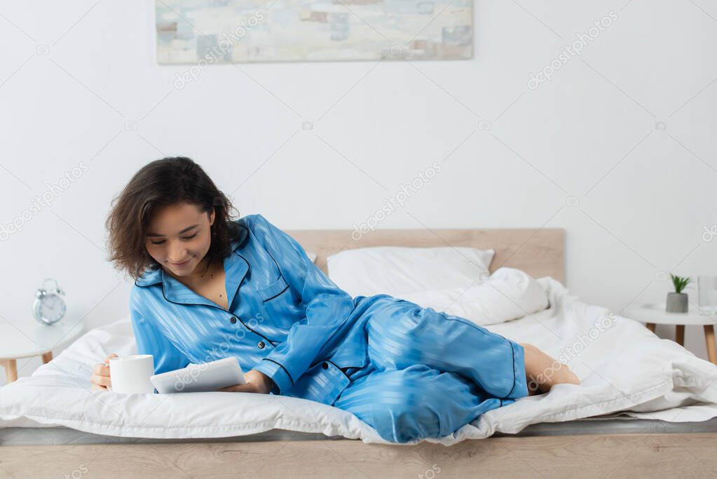 pleased woman in blue pajamas holding cup of coffee and using digital tablet in bedroom 
