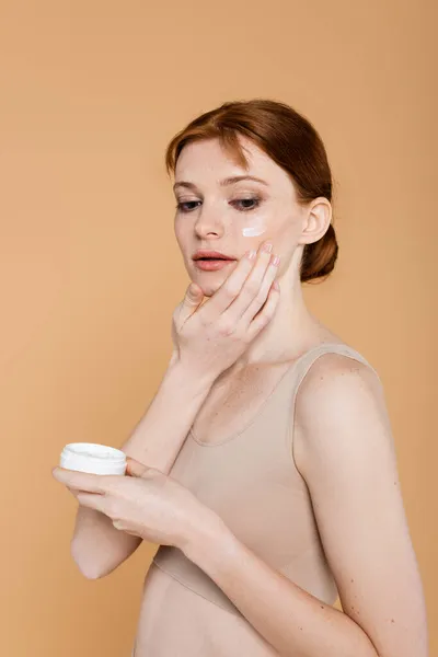 Freckled and redhead woman applying cream on cheek isolated on beige
