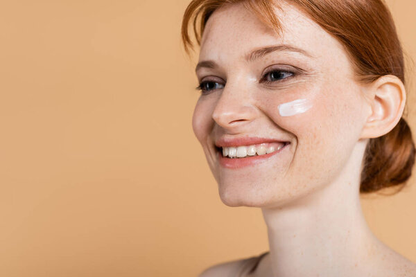 Smiling redhead woman with perfect skin and cream on face looking away isolated on beige 