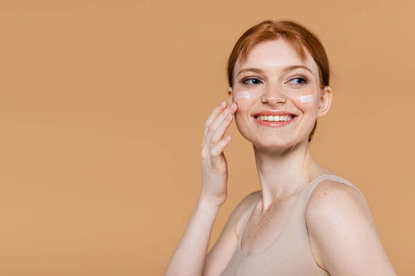 Positive red haired woman applying cosmetic cream on cheeks isolated on beige