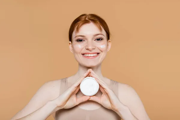 Uśmiechnięta Ruda Kobieta Trzymająca Krem Kosmetyczny Patrząca Kamerę Odizolowaną Beżu — Zdjęcie stockowe