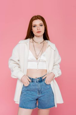 Pembe üzerine izole edilmiş şort ceplerinde el ele tutuşan ceketli genç kızıl saçlı kadın.