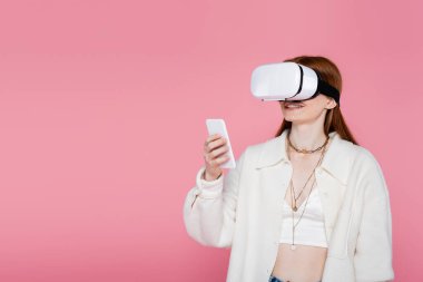 Sanal gerçeklik kulaklığı takmış gülümseyen stil sahibi bir kadın pembe üzerine izole edilmiş cep telefonu kullanıyor. 