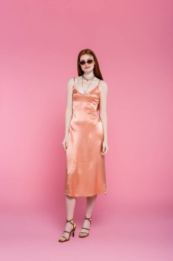 Full length of young red haired woman in sunglasses and dress standing on pink background  clipart