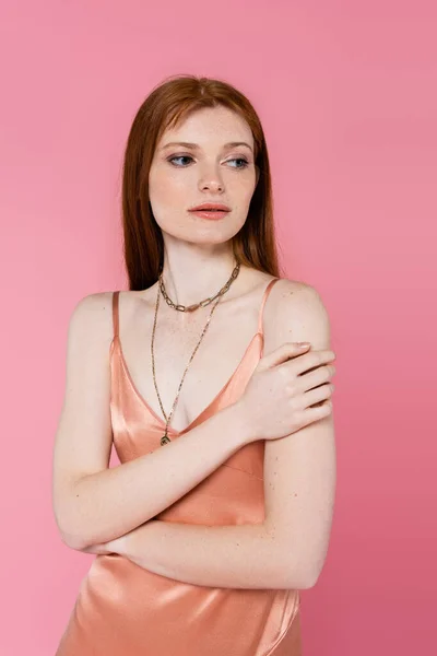 Young freckled and redhead woman in silk dress posing isolated on pink