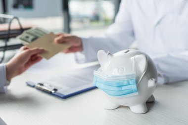 Tıbbi maskeli domuzcuk kumbarasının seçici odak noktası bulanık arka plan ve yolsuzlukla mücadele konseptiyle ilgili kadınlardan rüşvet alan kırpılmış bir doktorun yanı.