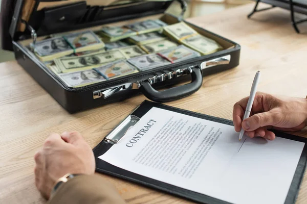 ドルと汚職防止の概念を持つぼやけたブリーフケースの近くにビジネスマンの署名契約のクロップドビュー — ストック写真