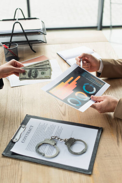 КИЕВ, УКРАИНА - 22 СЕНТЯБРЯ 2021: частичный взгляд бизнесменов с долларами и инфографикой возле наручников на контракты, антикоррупционная концепция
