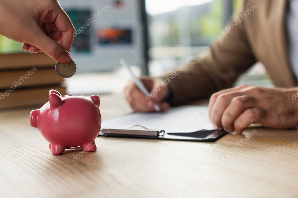 cropped view of woman holding coin near piggy bank while blurred businessman signing contract, anti-corruption concept