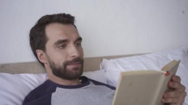 Yatakta kitap okurken gülümseyen sakallı adam. 