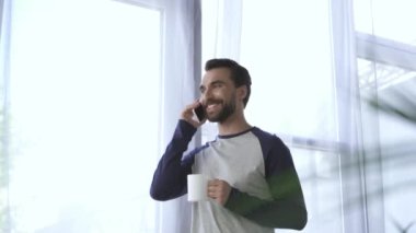 Elinde kupayla gülümseyen adam evde akıllı telefondan konuşuyor. 