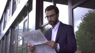 Gözlüklü bir işadamı bina yakınında gazete okuyor.