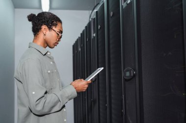 Veri merkezindeki sunucuların yanında dijital tabletli Afro-Amerikan programcının yan görüntüsü