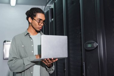 Gözlüklü Afro-Amerikan mühendis veri merkezindeki sunucuları kontrol ederken elinde laptop tutuyor.