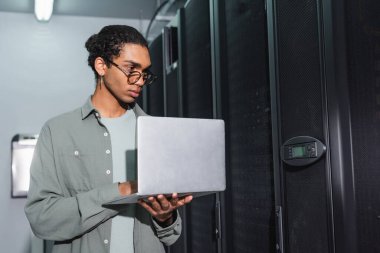 african american engineer in eyeglasses holding laptop while checking servers in data center clipart