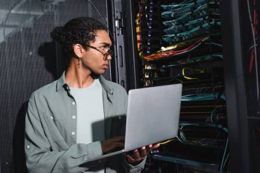 Afro-Amerikan mühendis sunucudaki kablolara bakarken dizüstü bilgisayar tutuyor.