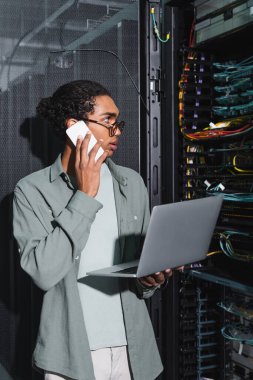 Diz üstü bilgisayarı olan Afrikalı Amerikalı teknisyen veri sunucusundaki sunucuları kontrol ederken akıllı telefondan konuşuyor