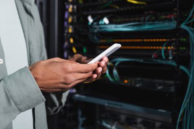 Veri merkezinde sunucu yakınında akıllı telefon kullanan Afro-Amerikan mühendis görüntüsü.