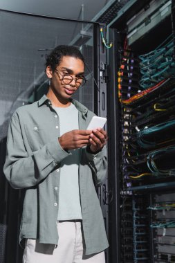 gülümseyen Afrikalı Amerikalı programcı veri merkezinde sunucuya tanı koyarken cep telefonu kullanıyor