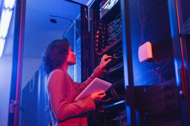 young technician holding digital tablet while checking wires in server in data center clipart