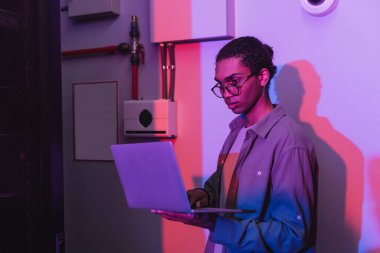 Gözlüklü genç Afrikalı Amerikalı programcı veri merkezinde, neon ışıkta dizüstü bilgisayar kullanıyor.
