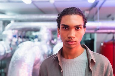 Piercingli genç Afro-Amerikan mühendis veri merkezi ekipman odasındaki kameraya bakıyor.