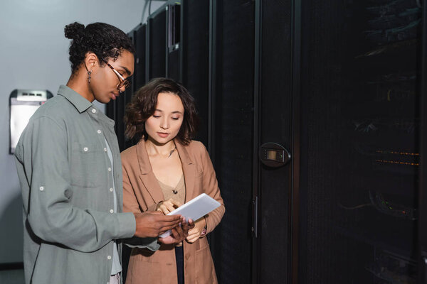 young programmer pointing at digital tablet while working with african american colleague in data center