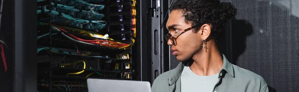 データセンター バナーで作業中にサーバー内のワイヤを見てラップトップを持つアフリカ系アメリカ人エンジニア — ストック写真