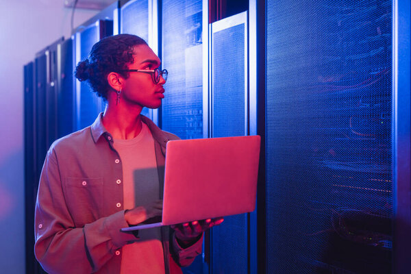 african american technician with laptop looking at servers in data center in neon light
