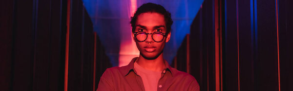 молодой африканский американский инженер смотрит на камеру в дата-центре с неоновым светом, баннер