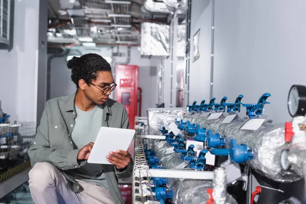 young afican american technician with digital tablet looking at cooling system of data center