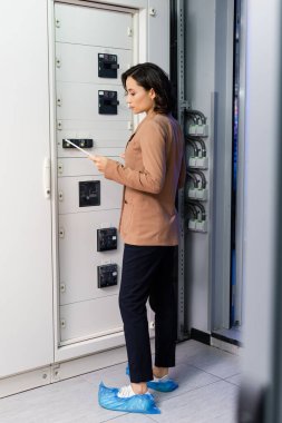 engineer looking at digital tablet while standing near switchboard in data center clipart