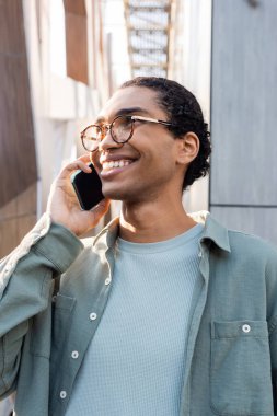 Gri gömlekli ve gözlüklü neşeli Afro-Amerikan adam şehir caddesinde akıllı telefondan konuşuyor.
