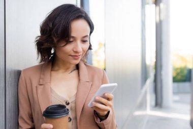 Dışarıdaki cep telefonuna bakmak için elinde kahve olan genç bir kadın.