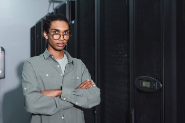 Gözlüklü Afrikalı Amerikalı programcı çapraz kollarla duruyor ve veri merkezindeki kameraya bakıyor.