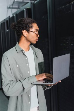 Genç Afrikalı Amerikalı programcı veri merkezinde sunucularla çalışırken dizüstü bilgisayar kullanıyor.