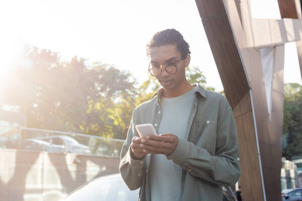 trendy african american man in eyeglasses messaging on mobile phone outdoors