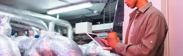 partial view of afican american engineer with digital tablet and paper cup in technical equipment room in data center, banner