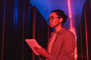 Dijital tabletli genç Afrikalı Amerikalı programcı neon ışıklı veri merkezinde sunucuların yanında çalışıyor.