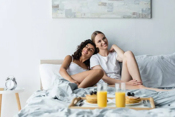 年轻快乐的女人靠在女友身边 吃着早餐躺在床上 — 图库照片