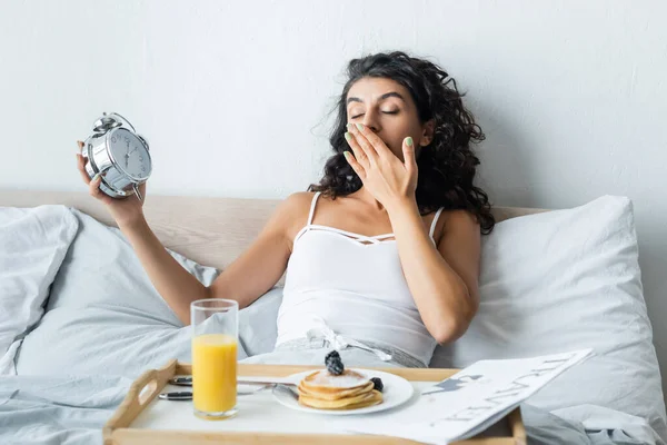 Mulher Sonolenta Cobrindo Boca Enquanto Bocejando Segurando Despertador — Fotografia de Stock