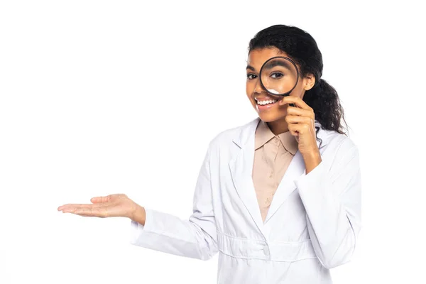 stock image Positive african american ophthalmologist holding magnifying glass and pointing with hand isolated on white 