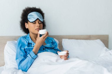 Afro-Amerikan kadın pijamalı ve uyku maskeli elinde konteynır ve yatakta kozmetik krem kokuyor. 