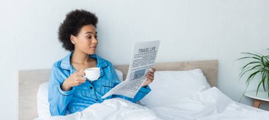 Afro-Amerikan kadın pijamalarıyla seyahat gazetesi okuyor. Elinde bir fincan kahve, pankart.