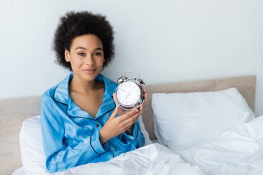 Pijamalı, gülümseyen Afro-Amerikan kadın yatakta çalar saat tutuyor.