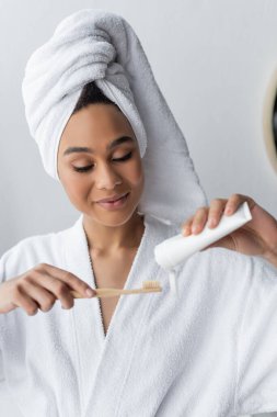 Bornoz ve havlu giymiş neşeli Afro-Amerikan kadın banyoda diş macunu ve diş fırçası tutuyor. 