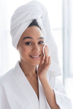Havlu giymiş neşeli Afro-Amerikalı kadın banyoda pamuk pedi ile yüzünü temizliyor. 