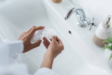 cropped view of african american woman holding bottle with cleanser and cotton pad near sink clipart