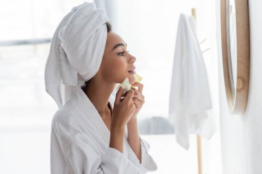 side view of young african american woman in white towel scrapping face with jade face scrapers and looking at mirror  clipart